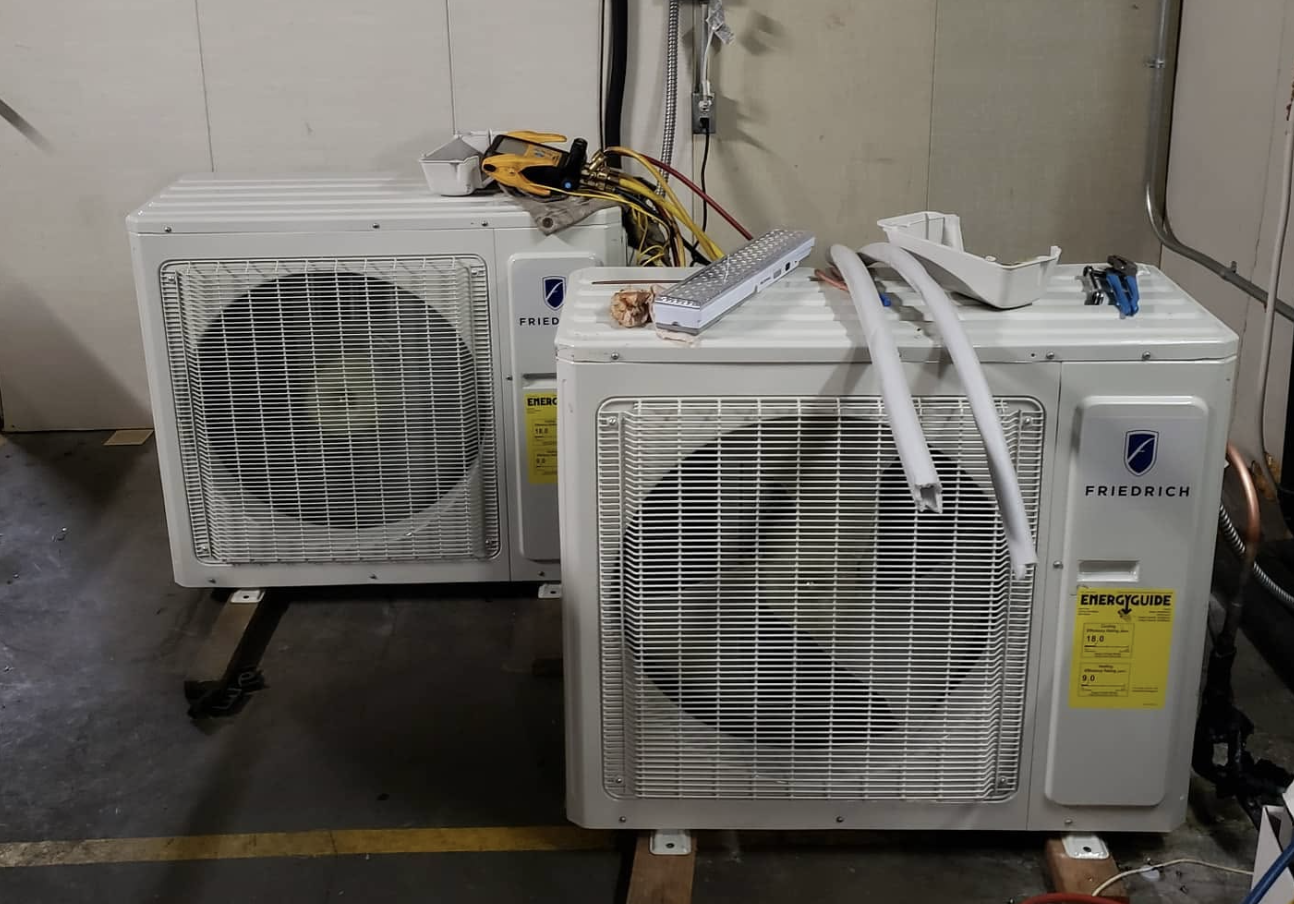 Two Friedrich ductless mini-split outdoor condenser units on timber supports in a workshop with manifold gauges during service